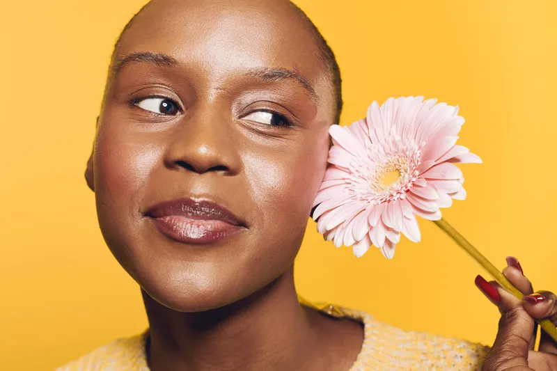 Close-up of a smiling young black woman with dark skin holding a pink gerbera flower near her face on a bright yellow background. Concept of beauty, happiness, and natural skincare.