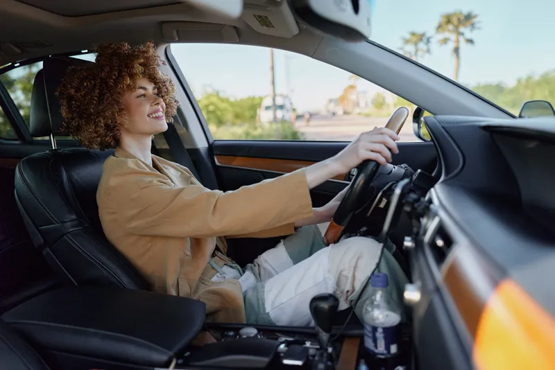 Young woman with curly hair drives a modern car, smiling and focused as she grips the steering wheel, captured in a candid roadside moment during daytime travel with warm, natural light.