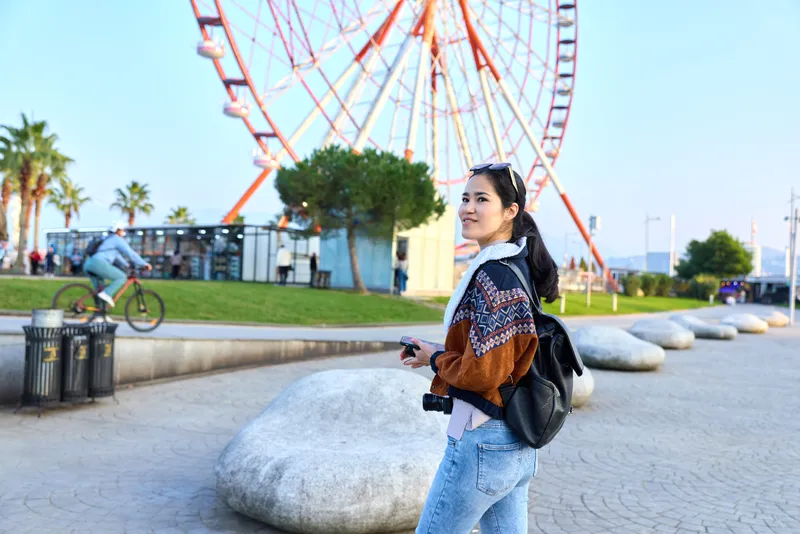 Woman with camera and backpack strolls a sunny boardwalk near a large ferris wheel, park benches and palm trees, capturing moments of travel, outdoor leisure, and casual fashion in a vibrant coastal