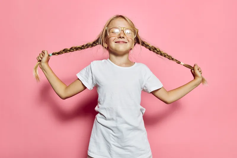 Happy girl in white t-shirt holding long braids with both hands smiling with closed eyes on pink background. Childhood fun and joy concept.
