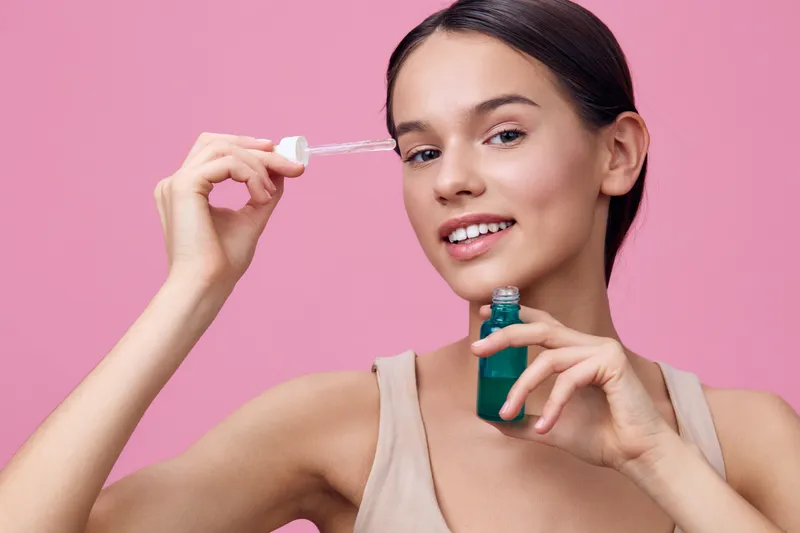 Young woman applying skincare serum with a dropper against a pink background, showcasing beauty and wellness in a fresh, vibrant style