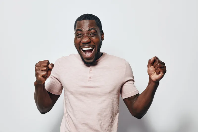 African American man with dark skin celebrating success with clenched fists and joyful expression wearing light pink shirt on white background.