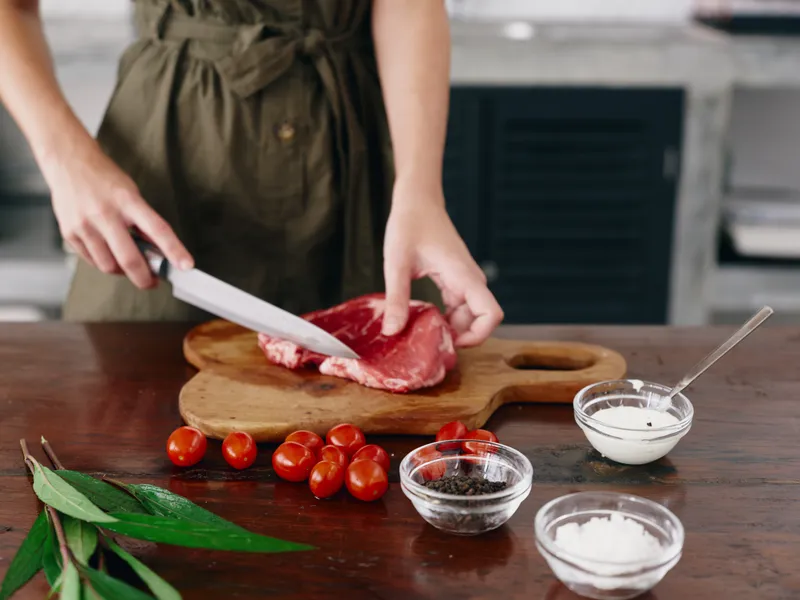 Женщина с ножом в руке нарезает стейк из мяса для жарки на кухне, на столе соль, перец и другие специи, красные помидоры черри и зелень, приготовление ужина