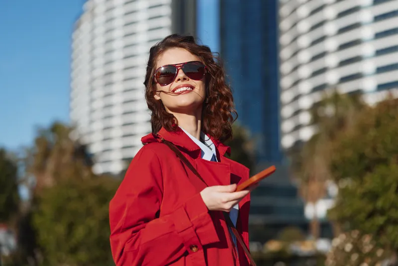 Smiling woman in red coat and sunglasses holds smartphone outdoors with modern buildings and clear sky in background, enjoying sunny day and urban lifestyle.