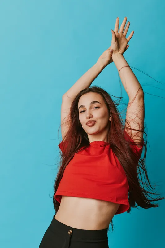Young woman with long hair wearing red crop top posing with raised arms against blue background, expressing joy and confidence with playful facial expression.