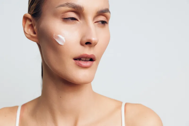 Young woman with clear skin applies facial cream on cheek with closed eyes wearing white tank top isolated on white background, skincare beauty treatment concept.