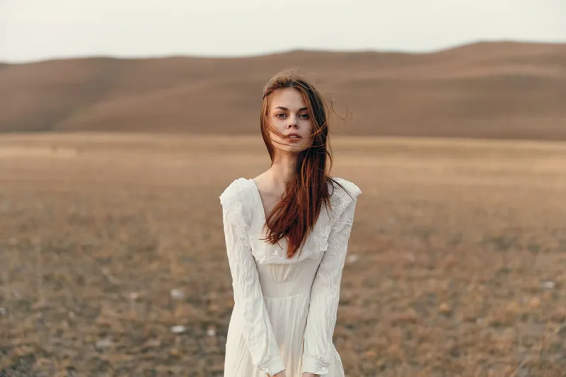 Женщина в белом платье стоит посреди открытого поля, уперев руки в бока, и наслаждается красотой природы и свободой