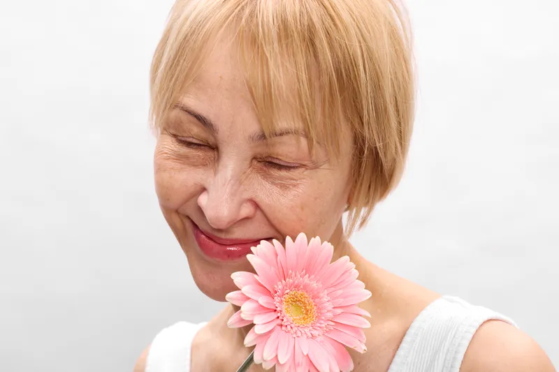Woman smiling, blonde short hair, holding a pink flower closeup, eyes closed, soft studio white background, spring wellness, relaxation, gentle portrait