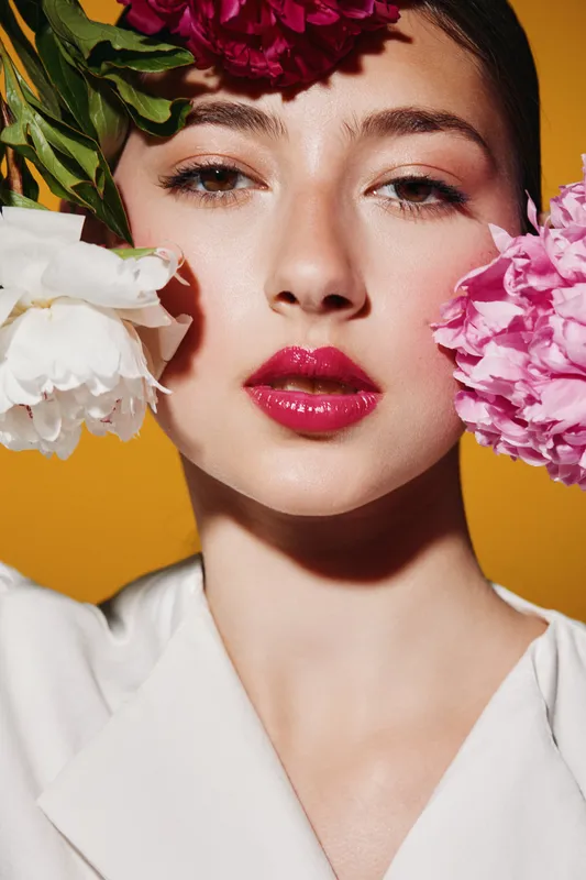 Close up portrait of young woman with vibrant flowers near face and bright glossy lips. Bold beauty fashion concept captured on vivid yellow background.