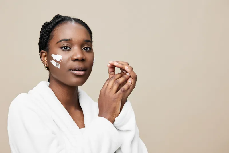 Young Black woman applying skincare cream on her face, showcasing healthy skin, in a light colored background, perfect for wellness and beauty concepts