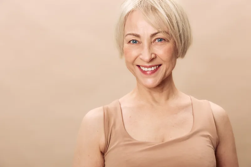 Smiling middle aged woman with short blonde hair and natural makeup wears beige tank top against light background. Concept for beauty, confidence, wellness, and positive aging.