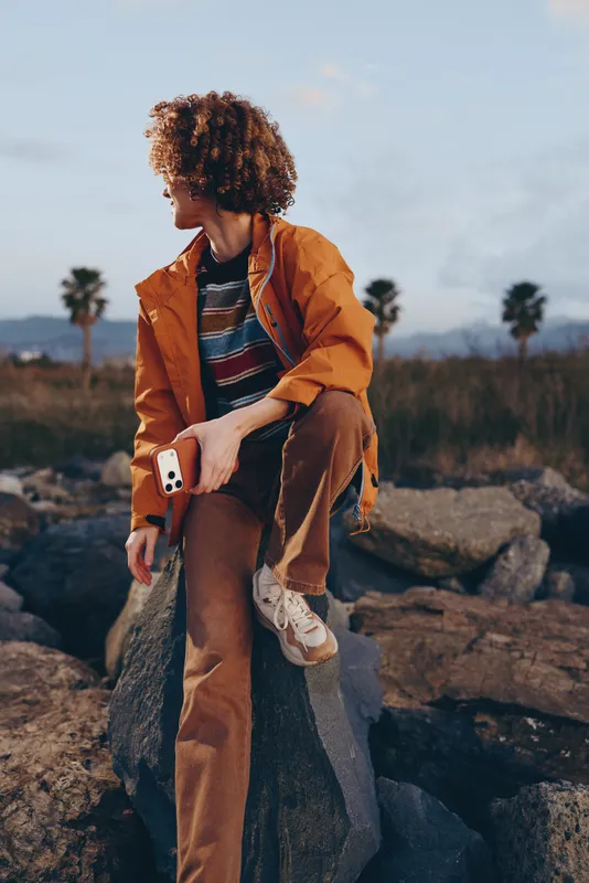 Woman lifestyle outdoors, sitting on rocks at golden hour, orange coat and curly hair, holding smartphone, thoughtful relaxed pose, desert landscape, soft breeze, natural light, travel mood