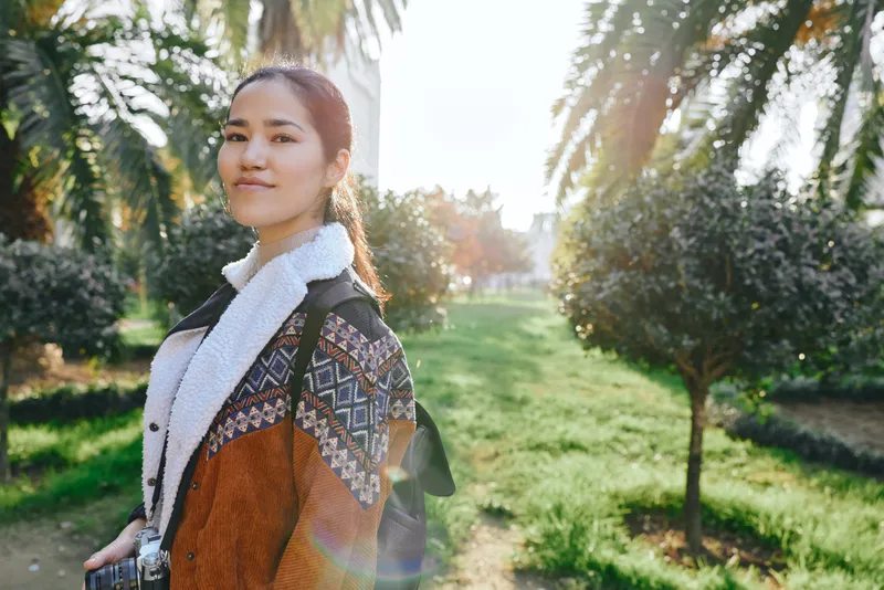 Woman wearing a patterned coat and scarf stands in a sunlit park, surrounded by greenery and trees, a calm outdoor fashion moment captured in natural daylight.