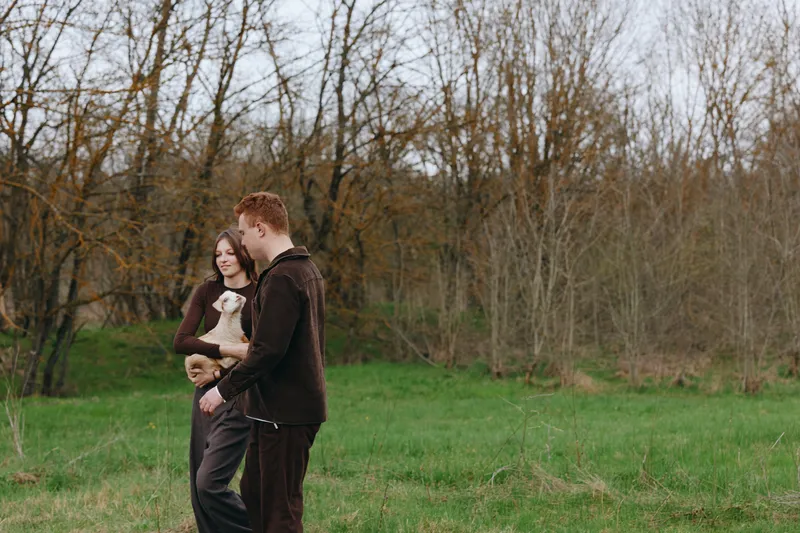 Cute couple outdoors, strolling with a playful goat kid, warm breeze, casual outfits, relaxed mood, lush meadow, sunlit backdrop, natural colors, candid lifestyle moment in open field