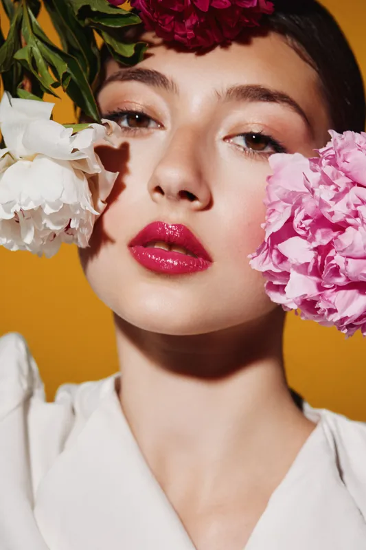 Closeup of a young woman with bright pink lipstick framed by vibrant flowers near face on an orange background. Concept of beauty, fashion, creativity, and floral portrait.