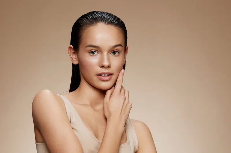 Natural beauty portrait of a young woman with smooth skin, wearing a neutral toned outfit, showcasing healthy skin and a serene expression against a soft beige background