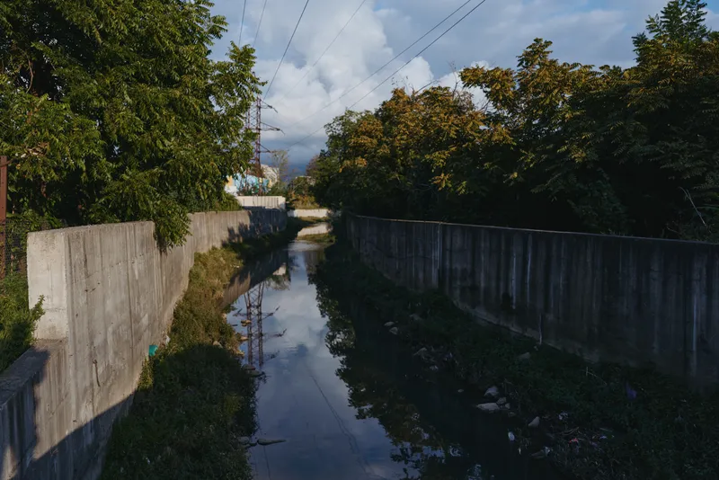 Concrete canal and urban drainage channel with reflective water under overcast sky, tree-lined concrete wall edges, empty industrial riverbed conveying stormwater and urban runoff infrastructure.