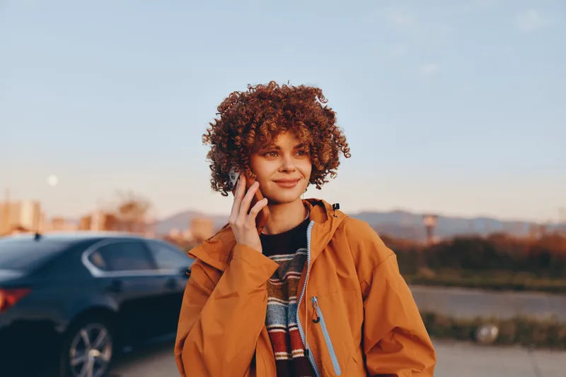 Woman lifestyle portrait outdoors in autumn, curly haired woman in brown jacket beside parked car, thoughtful smile during golden hour sunset, soft blurred city road background