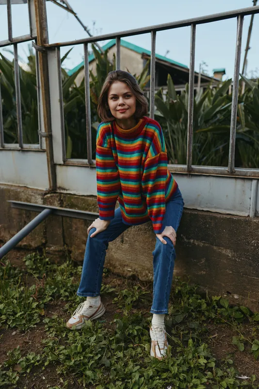 Woman in a colorful striped sweater and ripped jeans sitting on a metal railing outdoors, smiling and leaning forward in a casual outfit with white sneakers in tropical outdoor garden.