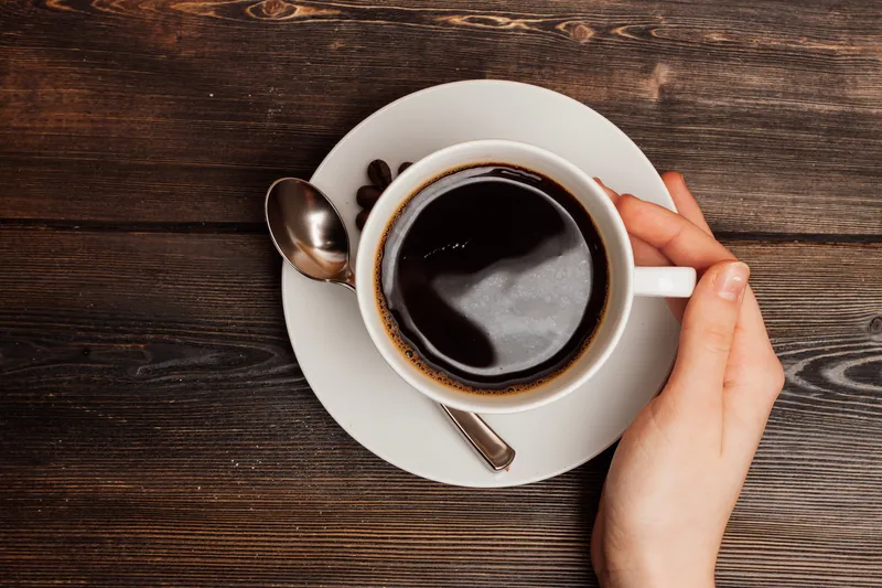 Кружка с горячим кофе стоит на деревянном столе, рядом лежит остаток завтрака или перекуса