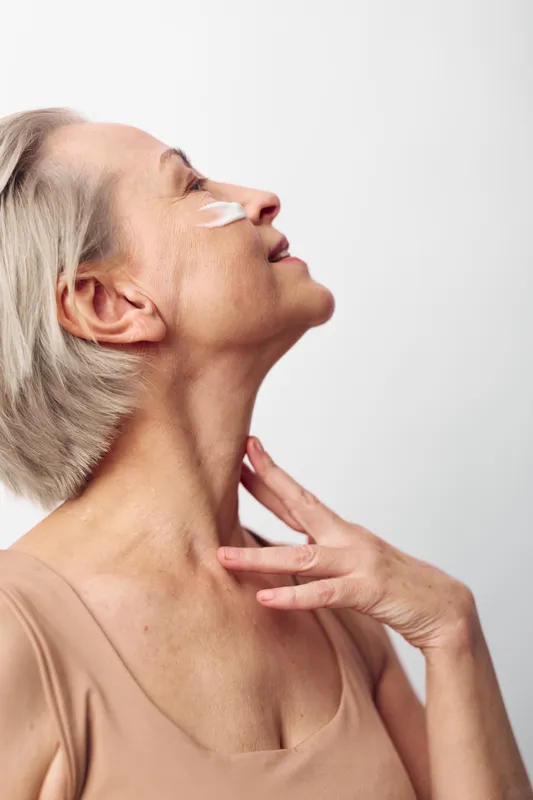 Mature woman with short grey hair applies cream on her face with gentle touch, eyes closed in peaceful moment. Concept of skincare, aging, self-care, and natural beauty on white background.