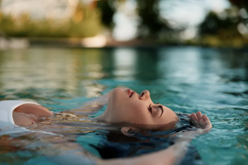 Relaxation on a sunlit day as a person floats on her back in a clear pool, eyes closed, peaceful expression, warm breeze, soft reflections, vibrant water, casual summer mood