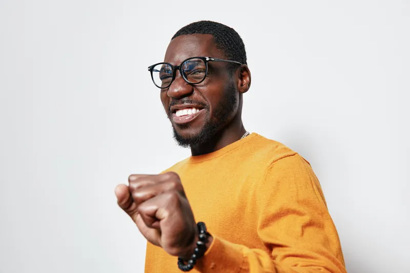 African man with dark skin wearing glasses and orange sweater smiling and celebrating success with fist pump on white background. Positive emotion and casual style portrait.