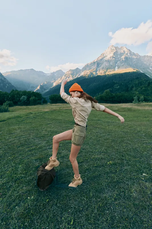 Young woman in an orange beanie and khaki shorts joyfully jumps on a backpack in a green meadow with mountains in the background during a sunny day.