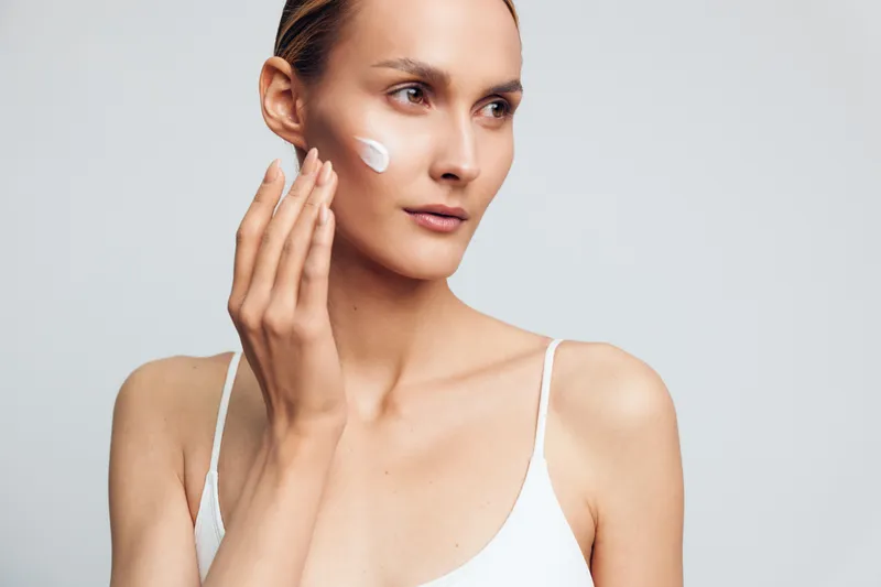Young woman applies face cream with fingers focusing on skincare routine with natural clear skin wearing white top isolated on light gray background.