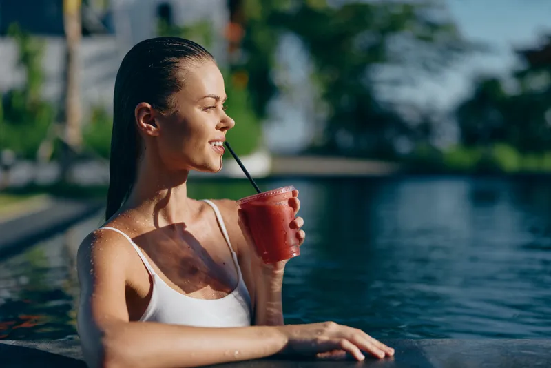Young woman with wet hair relaxing in pool drinking red smoothie through straw, enjoying summer lifestyle outdoors on sunny day with blurred natural background and calm water.