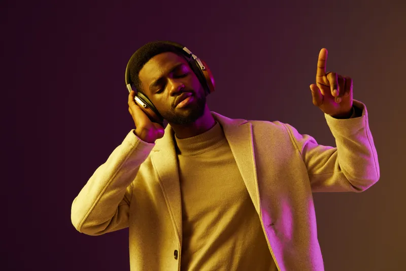 Young Black man enjoying music with headphones, smiling and dancing in stylish fashion against a vibrant purple background