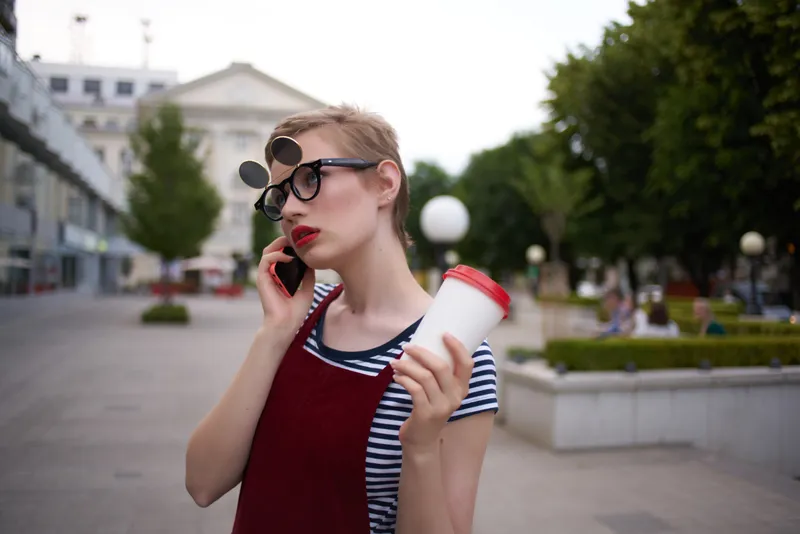 Женщина в очках на улице разговаривает по телефону, держа в руке стакан с напитком