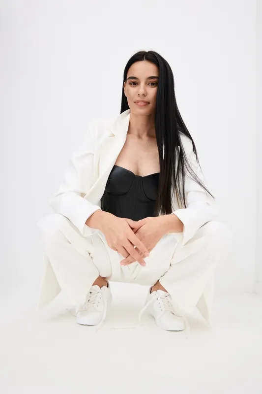 Woman with long black hair wearing white suit and black top sitting on white floor with confident and calm expression in minimalistic bright studio setting.
