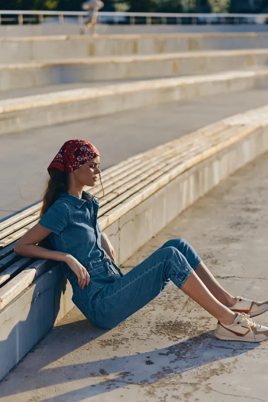 Lifestyle woman in modern boho-western denim street style, sitting relaxed outdoors wearing a red bandana and casual white sandals, film color tones, warm sunlight.