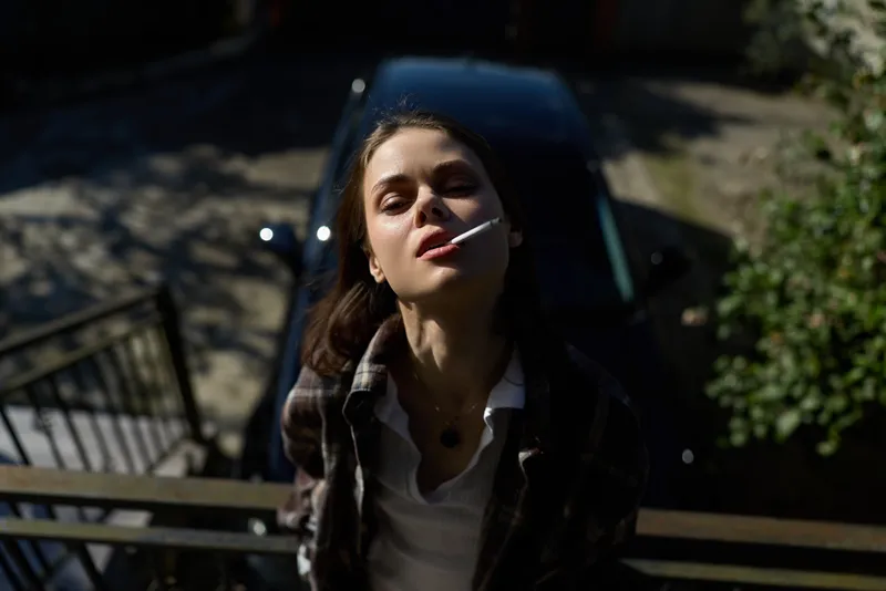 Cheerful woman outdoors, smoking on sunlit stairs, casual jacket, relaxed posture, natural light, soft breeze, urban greenery background, candid moment