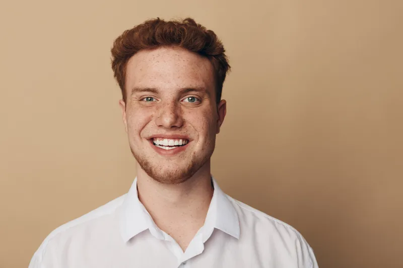 Smiling young man with red hair and freckles wears white shirt against beige background. Portrait of happy male model with natural skin and casual style. Concept of youth and positivity.