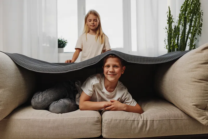 Happy children playing hide and seek under a blanket on a sofa in bright living room. Concept of family fun, childhood joy, and indoor activity with natural light atmosphere.