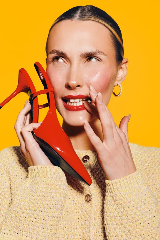 Woman with slicked hair holds red high heel shoe near face, wearing beige sweater and gold hoop earrings. Bright yellow background creates vibrant fashion and beauty concept with confident mood.