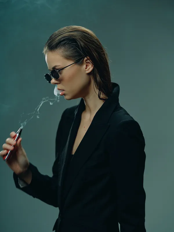 Stylish woman smoking vape in black jacket and sunglasses, modern urban fashion with moody atmosphere and cool attitude.