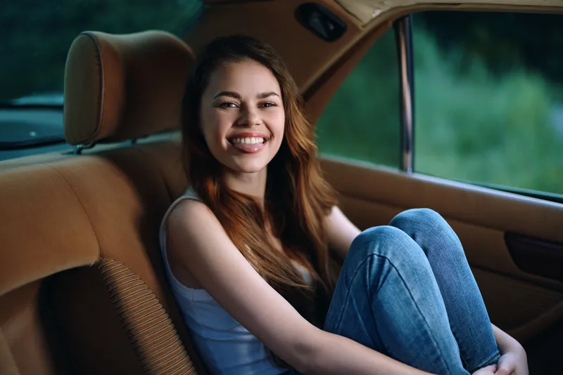 Happy young woman sitting in the backseat of a vintage car, wearing casual clothes, smiling joyfully against a green blurred background.
