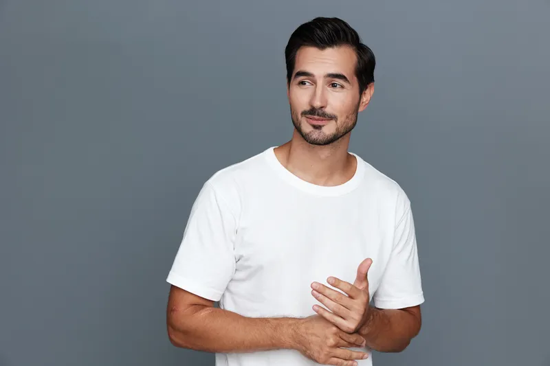 Smiling man wearing a casual white t-shirt stands against a gray background, expressing a thoughtful demeanor with subtle confidence. Portrait lifestyle concept.
