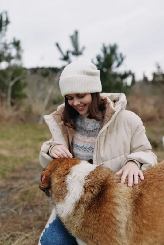 Woman in a warm coat and beanie smiles while stroking a large dog outdoors, embracing nature on a chilly day in an open park setting with trees, soft light and a joyful moment