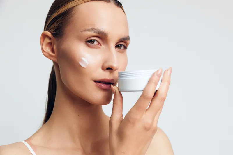 Young woman with blonde hair applies white face cream on her cheek while holding an open jar against a plain light background, demonstrating skincare and self-care routine.