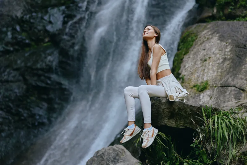 Woman sitting peacefully near a large waterfall surrounded by rocks and greenery, wearing white casual clothes and enjoying nature in a serene outdoor setting.