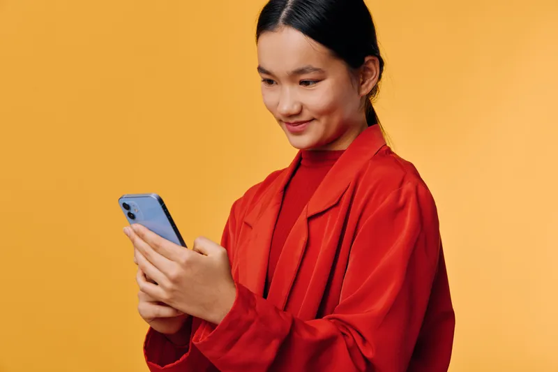 Young woman in red jacket smiling and using smartphone with both hands on orange background. Portrait, technology, communication, casual, connection, digital device.