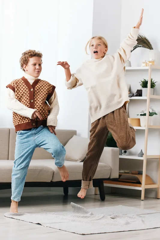 Two playful children jump indoors on a soft rug, capturing spontaneous joy and energy as one kid raises a hand mid air in a bright, cozy living room.