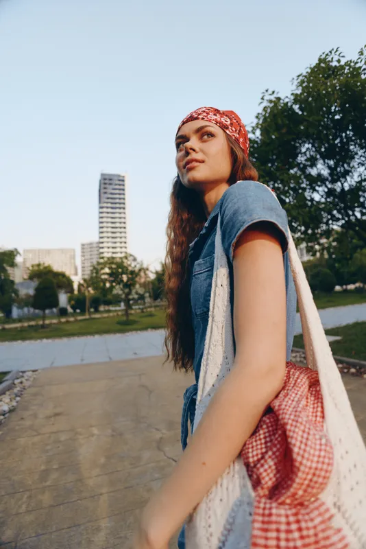young woman with red bandana and casual denim outfit walking outdoors in urban park on sunny day, carrying a red checkered bag over shoulder