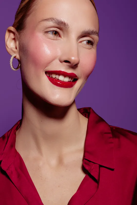 Close-up of a smiling woman with red lipstick and hoop earrings wearing a red blouse. She looks away from the camera against a purple background. Concept of beauty and fashion.