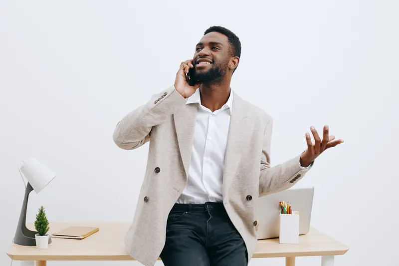 Confident man in beige blazer and white shirt talking on mobile phone, smiling happily while standing near modern desk with office supplies, on plain white background. Business communication concept.
