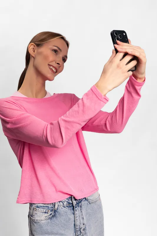 Young woman in pink sweater takes a selfie with smartphone on white background. She smiles naturally, holding the phone with both hands in casual modern outfit.